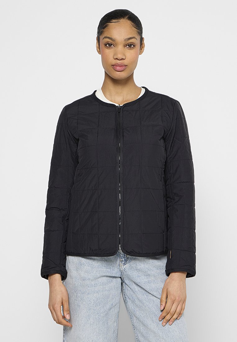 Giovane donna che indossa una giacca nera trapuntata aderente con chiusura a zip e jeans azzurri, in piedi di fronte a uno sfondo bianco uniforme.