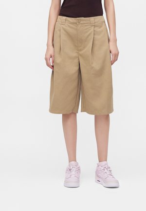 Personne portant un short beige ample jusqu'aux genoux, des baskets rose clair et un haut noir sans manches, debout sur un fond blanc.