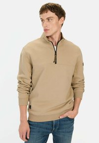 Beige sweatshirt met ritssluiting en lange mouwen, met elastische manchetten, voorzien van een gestructureerde kraag en een contrasterende ritshendel. Vergezeld van blauwe jeans.