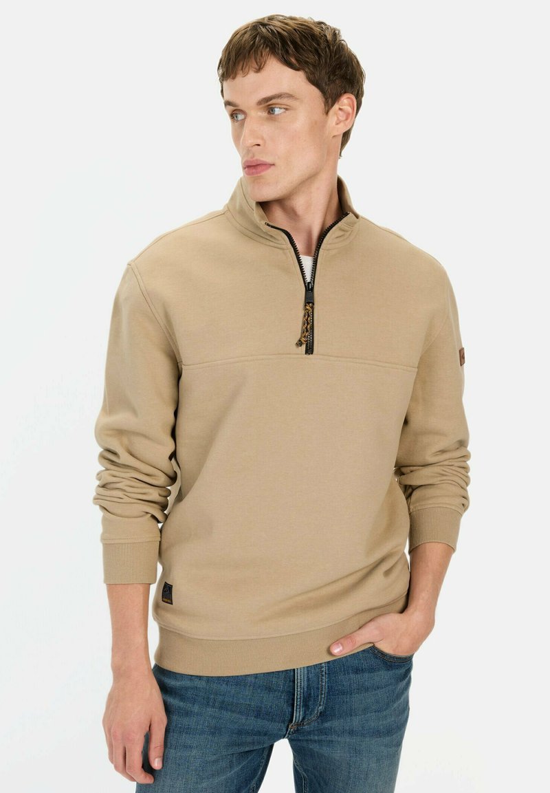 Beige Zip-Hals-Sweatshirt mit langen Ärmeln und elastischen Bündchen, ausgestattet mit einem strukturierten Kragen und einem kontrastierenden Reißverschluss. Dazu werden blaue Jeans getragen.