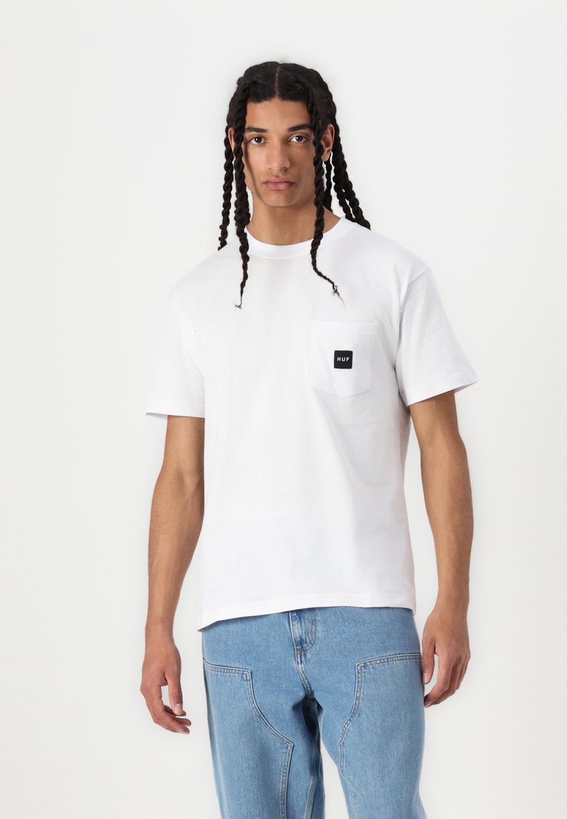 Tricou alb din bumbac cu guler rotund și mâneci scurte, dotat cu un buzunar pe pieptul stâng, având o etichetă neagră. Asortat cu blugi subțiri de culoare bleu.
