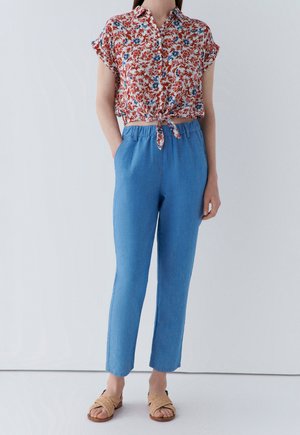 Bluse mit floralem Muster und kurzen Ärmeln mit Bindeband vorne; hellblaue, elastische Hose; beige, geflochtene Sandalen; glatte Stofftextur.
