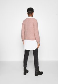 Różowy sweter na drutach z długimi rękawami, noszony na białej koszuli z kołnierzykiem, zestawiony z ciemnymi dżinsami i czarnymi botkami za kostkę.