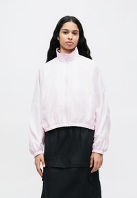 Ανοιχτό ροζ ζακέτα με φερμουάρ, ελαστικά μανσέτες και μέση, με ψηλό γιακά και λεία, γυαλιστερή υφή. Φοράται πάνω από μια μαύρη φούστα.