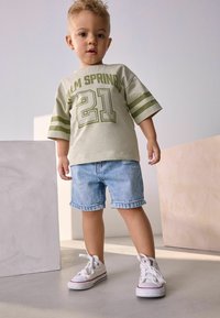 Camisa de manga corta verde claro con "Palm Springs 21" en verde oscuro, combinada con pantalones cortos de mezclilla. Zapatillas blancas completan el conjunto.