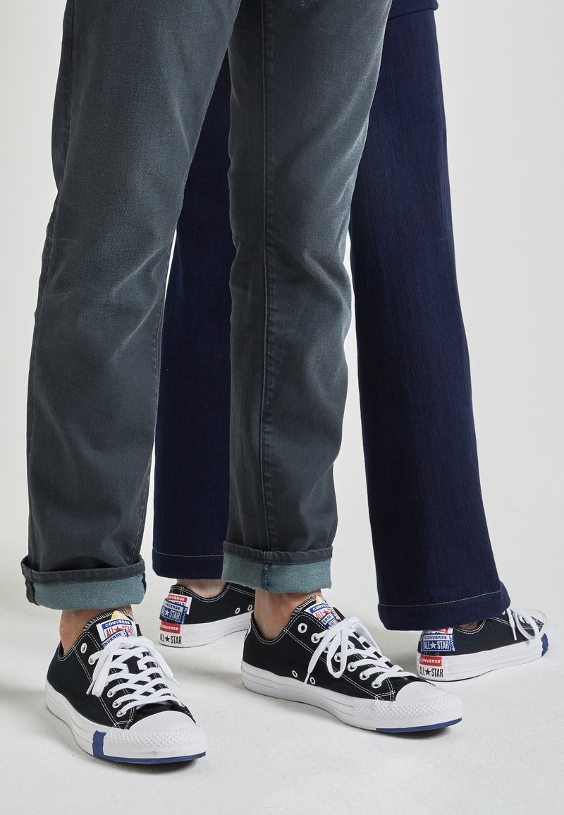 Svarta canvas-sneakers med vita gummisulor och snören. Har logotyp-lappar på hälen och flerfärgade detaljer, matchat med mörka jeans.