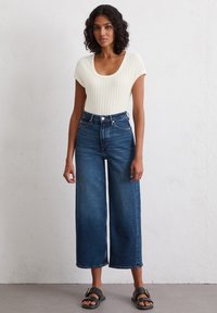 Helle cremefarbene, gerippte Kurzarmbodysuit, hochtaillierte dunkelblaue weite Jeans und schwarze Sandalen mit goldenen Akzenten. Einfaches Design und Passform.