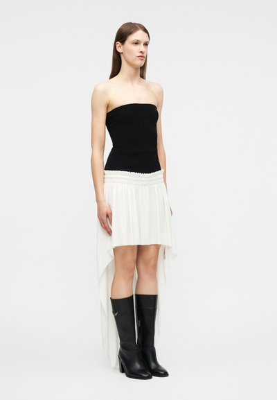 Γυναίκα με μαύρο τοπ χωρίς τιράντες, λευκή φούστα υψηλή-χαμηλή με σούρες στη μέση και μαύρες μπότες με τακούνι μέχρι το γόνατο, στέκεται μπροστά σε απλό φόντο.