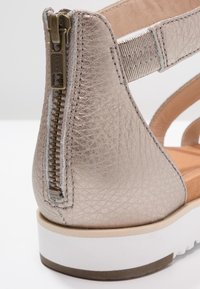 Zbliżenie na metaliczne beżowe skórzane sandały na obcasie z tylnym zamkiem błyskawicznym, elastycznym paskiem i grubą białą podeszwą.