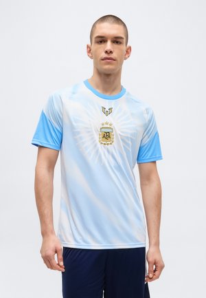 Jeune homme debout portant le maillot de football de l'Argentine bleu clair et blanc avec l'écusson AFA et un short sombre sur un fond uni.
