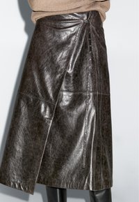 Dunkelbrauner Leder-Midirock mit Wickeldesign und Ösenriemen-Detail, kombiniert mit schwarzen Stiefeln und beigem Pullover.