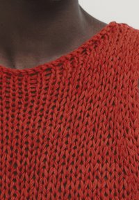 Punainen neulepusero löysällä kudonnalla, jossa on avara kaula-aukko ja tekstuuri. Materiaali näyttää pehmeältä ja kevyeltä.