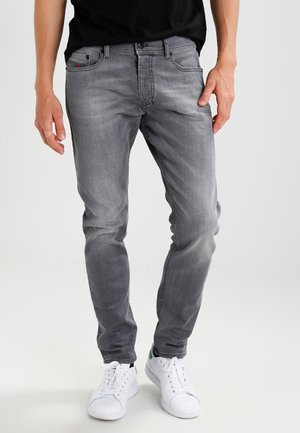 Personne portant un jean gris délavé et des baskets blanches, debout devant un fond clair uni.