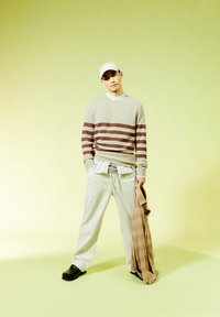 Maglione a righe beige con strisce marroni; pantaloni leggeri grigi larghi; zoccoli neri; tiene una sciarpa a scacchi beige; sfondo bianco uniforme.