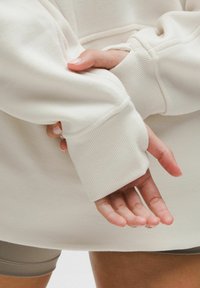 Roke križi za hrbtom, oblečen v oversize svetlo bež rebrast pulover in taupe kratke hlače, prsti sproščeni in vidni.
