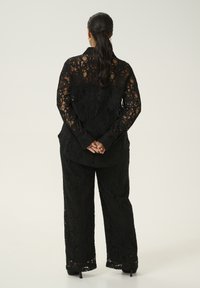 Zwart kanten twee-delige pak met lange mouwen, losse broek en ingewikkelde bloemenpatronen, met doorschijnende textuur en bijpassende zwarte hakken.