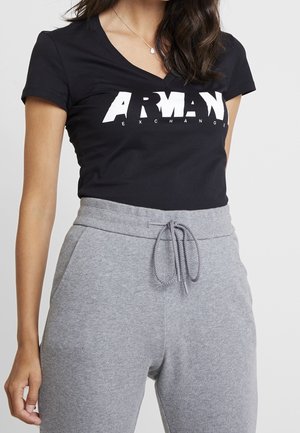 Mujer con camiseta de cuello en V negra de Armani Exchange y pantalones deportivos gris claro con cordón, mostrando el torso y el brazo derecho.