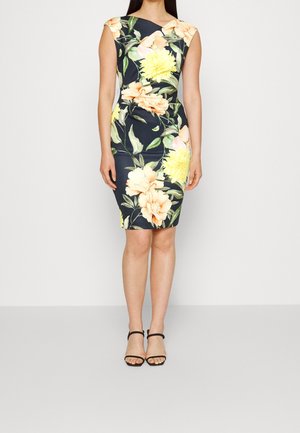 Femme portant une robe noire sans manches mi-longue aux genoux avec de grands imprimés floraux jaunes et pêche et des sandales noires à brides.