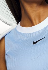 Jasnoniebieski sportowy top bez rękawów z białym kołnierzykiem, wykonany z teksturowanej tkaniny, z czarnym logo Nike na piersi.