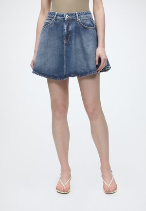 Femme portant une jupe en jean bleue avec des poches avant, associée à des sandales beige type tongs, debout sur un fond clair uni.