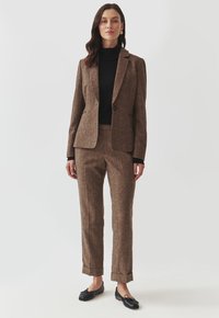 Blazer din lână texturată de culoare maro și pantaloni asortați, perecheți cu un pulover negru cu guler înalt. Pantofii loafer negri cu accente sub formă de fundiță completează ținuta.