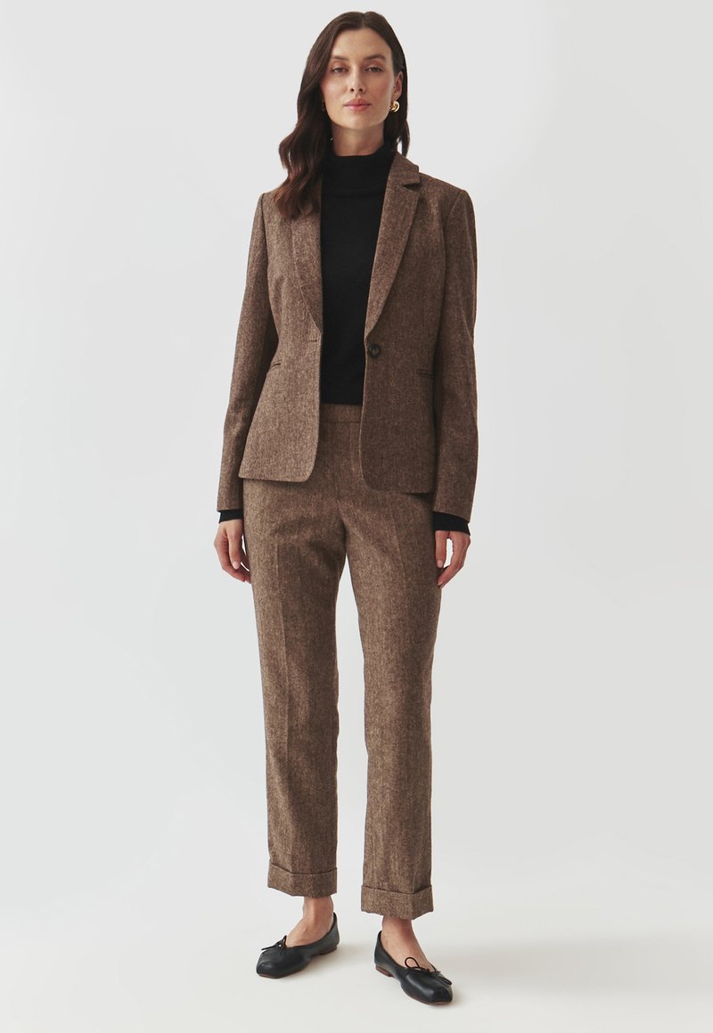 Blazer din lână texturată de culoare maro și pantaloni asortați, perecheți cu un pulover negru cu guler înalt. Pantofii loafer negri cu accente sub formă de fundiță completează ținuta.