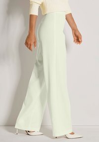 Vrouw in een hoge taille, met wijde crèmekleurige broek en witte puntige hakken, die zijwaarts tegen een lichte achtergrond staat.