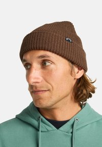Gorra de lana marrón con textura acanalada y un pequeño logo de la marca. Se ajusta cómodamente alrededor de la cabeza, diseñada para mantener el calor y para un uso casual.