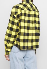 Person wearing a yellow and black checkered long-sleeve shirt and light blue jeans, shown from the back against a plain background.