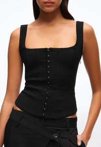 Nainen, joka käyttää istuvaa mustaa korsettityylistä toppia, jossa on koukku- ja silmukkakiinnitys edessä sekä leveät olkaimet, yhdistettynä mustiin housuihin, valkoista taustaa vasten.