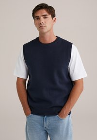 Mornarska pulover jakna s okruglim izrezom i bez rukava, uparena s bijelom majicom kratkih rukava, nosi se preko svijetloplavih traperica.