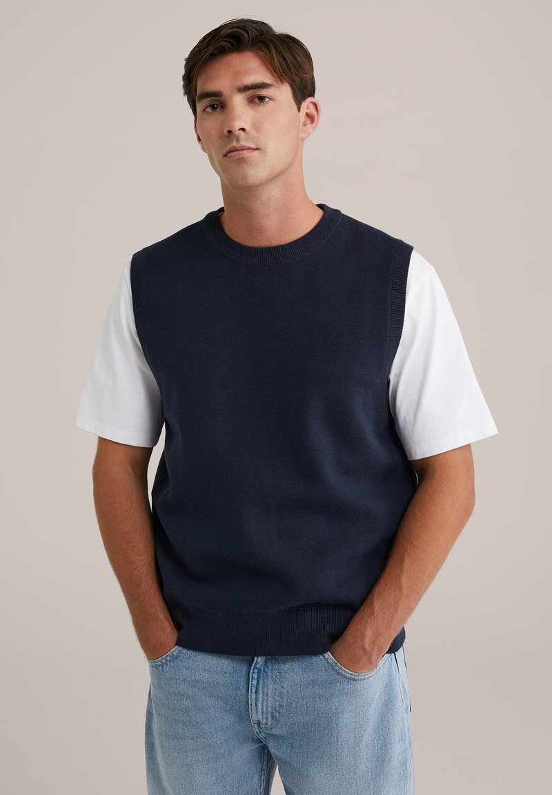 Mornarska pulover jakna s okruglim izrezom i bez rukava, uparena s bijelom majicom kratkih rukava, nosi se preko svijetloplavih traperica.