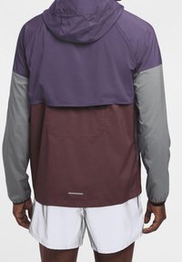 Mann in einer Kapuzenwindjacke mit lila, kastanienbraunen und grauen Farbblöcken, kombiniert mit weißen Sportshorts, von hinten betrachtet.