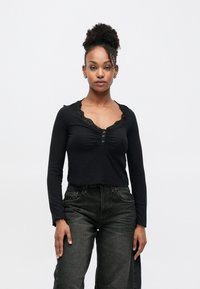 Černý top s dlouhým rukávem, výstřihem do V s krajkou, nařaseným předním dílem a knoflíkovými detaily. Kombinován s tmavými džínami s vysokým pasem.
