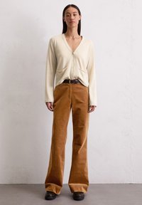 Eine Frau steht vor einer weißen Wand und trägt eine beige, knöpfbare Strickjacke, braune, hoch taillierte Cordhosen mit Gürtel und dunkle Schuhe.