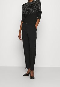 Pull noir avec un motif perlé au niveau de l'encolure, associé à un pantalon noir ajusté avec une fente discrète. Des chaussures noires en cuir verni complètent la tenue.