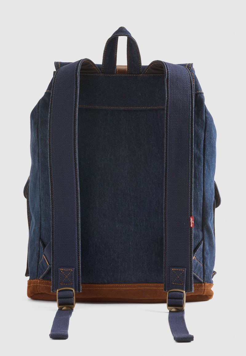 Zaino in denim con corpo blu scuro, fondo in suede marrone, spallacci imbottiti blu navy e maniglia superiore. Presenta cuciture a contrasto e dettagli in metallo.