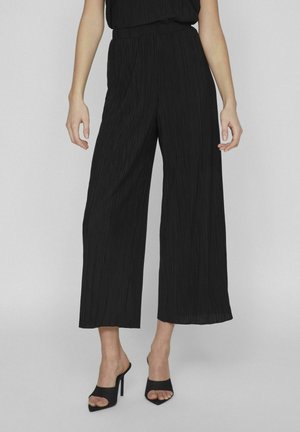 Mujer con pantalones negros anchos, plisados y recortados y zuecos negros de tacón alto y puntera abierta, de pie contra un fondo liso.