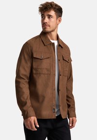Braune, weiche Stoffjacke mit zwei Brusttaschen, durchgehender Knopfleiste und schwarzen Knöpfen; lange Ärmel und gerade geschnitten. Über einem weißen Hemd getragen.