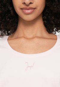 Lähivõte naeratusest inimesest lokkis juustega, kes kannab heledat roosat pluusi, mille kaeluse lähedal on diskreetne logo.