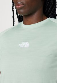 Camiseta de manga corta de color verde claro, confeccionada en un tejido texturizado, con un logo bordado en blanco en el pecho y un cuello redondo.