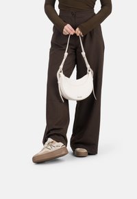 Une personne portant un haut marron ajusté tient un sac à main blanc, vêtue de larges pantalons marron et de baskets beiges, se tenant devant un fond uni.