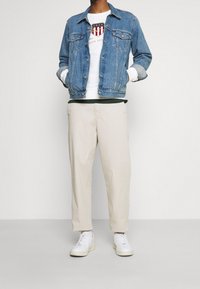 Jeansjacke in Hellblau, weißer Sweatshirt mit Logo, beigefarbene Hose und weiße Sneaker. Lässiges Outfit mit geschichteten Texturen.