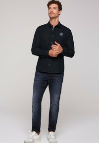 Camisa negra de manga larga con cuello abotonado y logotipo bordado, combinada con jeans oscuros y zapatillas blancas. Diseño simple y ajustado.