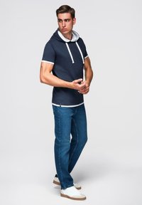 Námořnická krátká kapuce s bílými tkaničkami, spárovaná s denimovými džíny. Model stojí v neutrální póze, která ukazuje střih oblečení.