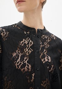 Gros plan d'une personne portant un haut noir en dentelle à boutons avec des motifs floraux et un col lisse sur fond blanc.