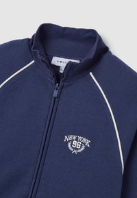 Giacca zip-up navy in cotone, con piping crema, colletto alto e logo ricamato "NEW YORK 96" con accenti di alloro.