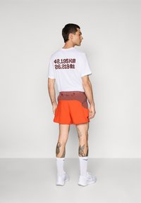 Mann mit weißen Turnschuhen, weißen Socken, roten Laufshorts und einem weißen T-Shirt, auf dessen Rücken "42,195 km 26,219 mi" steht, steht seitlich.