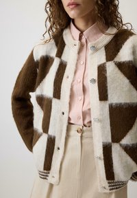 Kobieta nosząca brązowy i biały sweter w geometryczny wzór, założony na jasnoróżową koszulę z guzikami i beżowe spodnie z wysokim stanem.