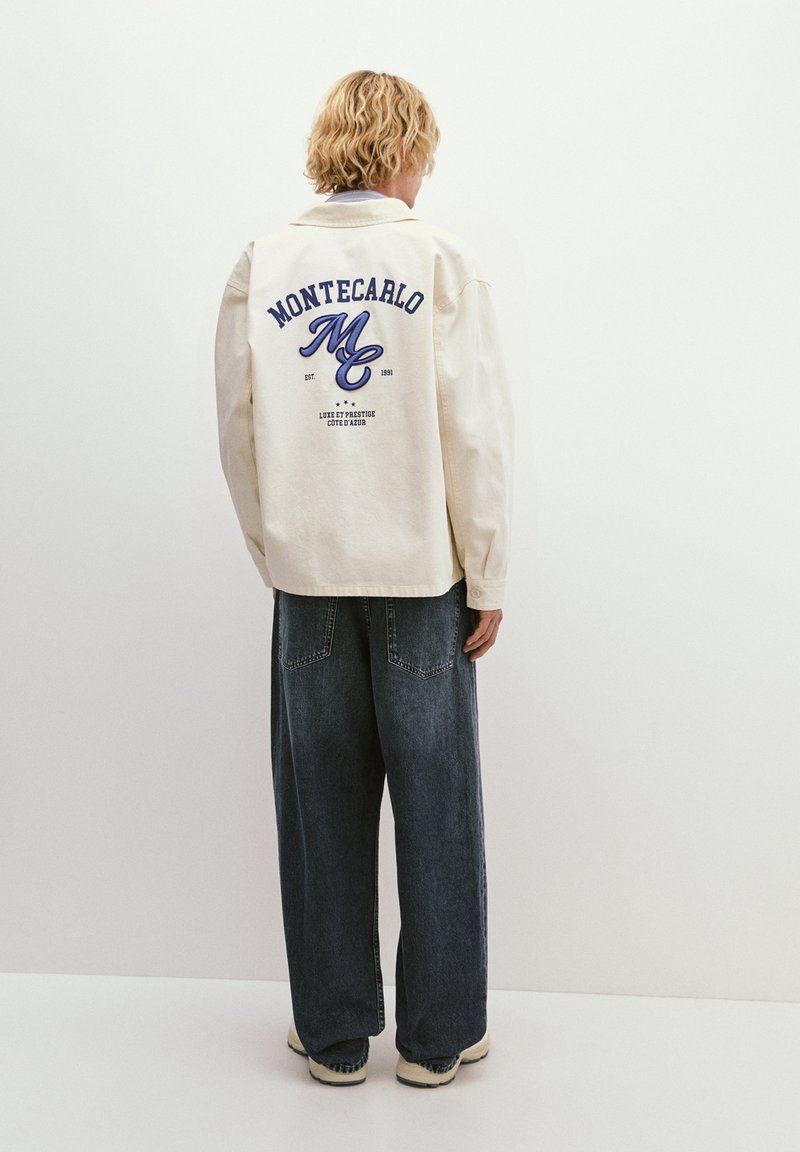 Cream-colored jacket with blue embroidered "MONTECARLO" text on the back, worn over dark blue wide-leg jeans and beige sneakers.